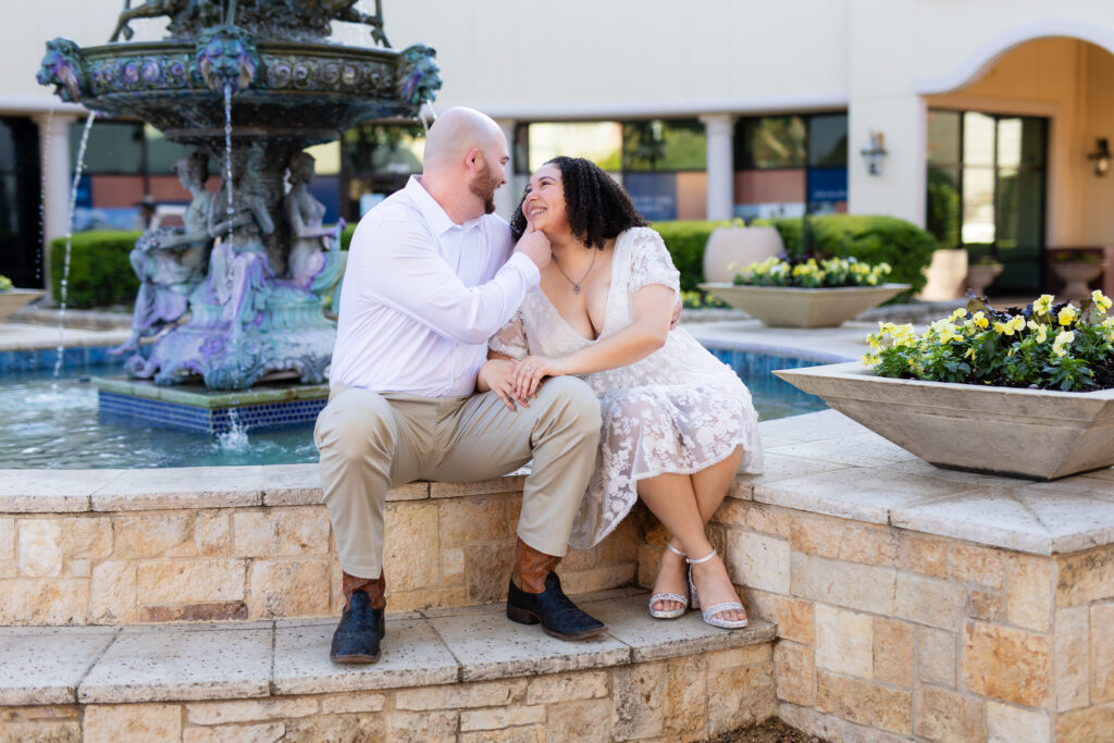 engaged couple wearing all white sitting on fountain ledge while man pulls woman's face in for kiss smiling