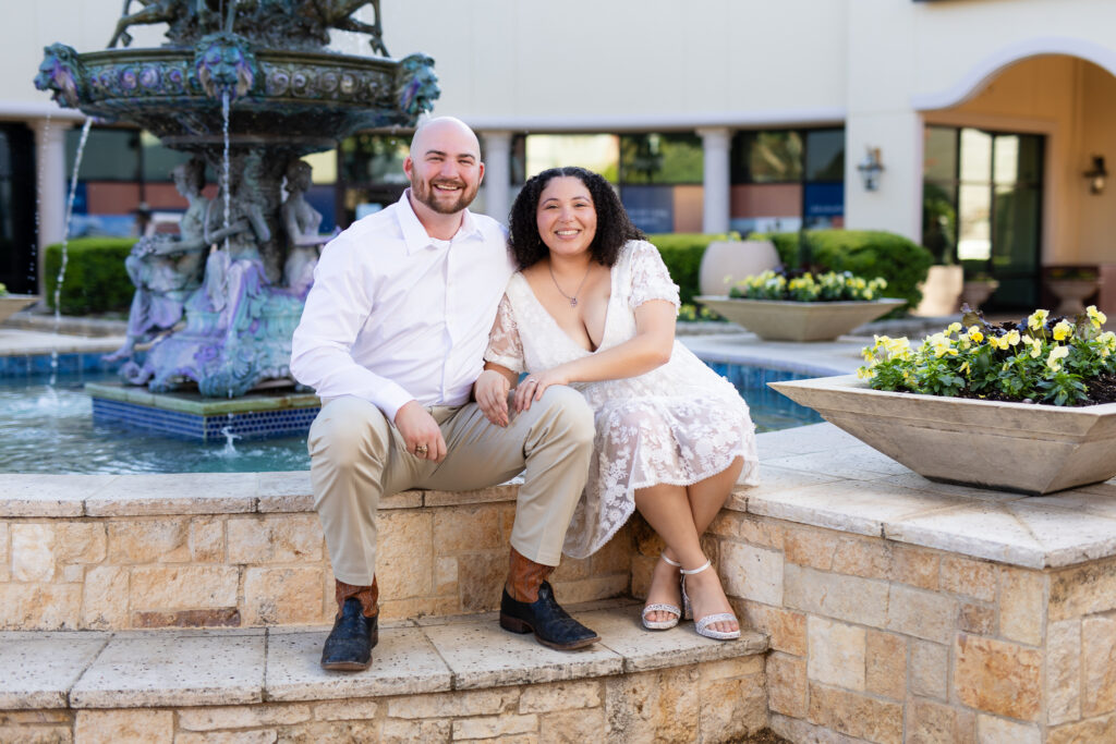 engaged couple sitting on fountain in Rockwall Texas smiling during engagement session