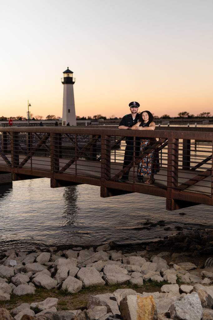 engaged couple standing on bridge with lighthouse behind them during sunset in Rockwall