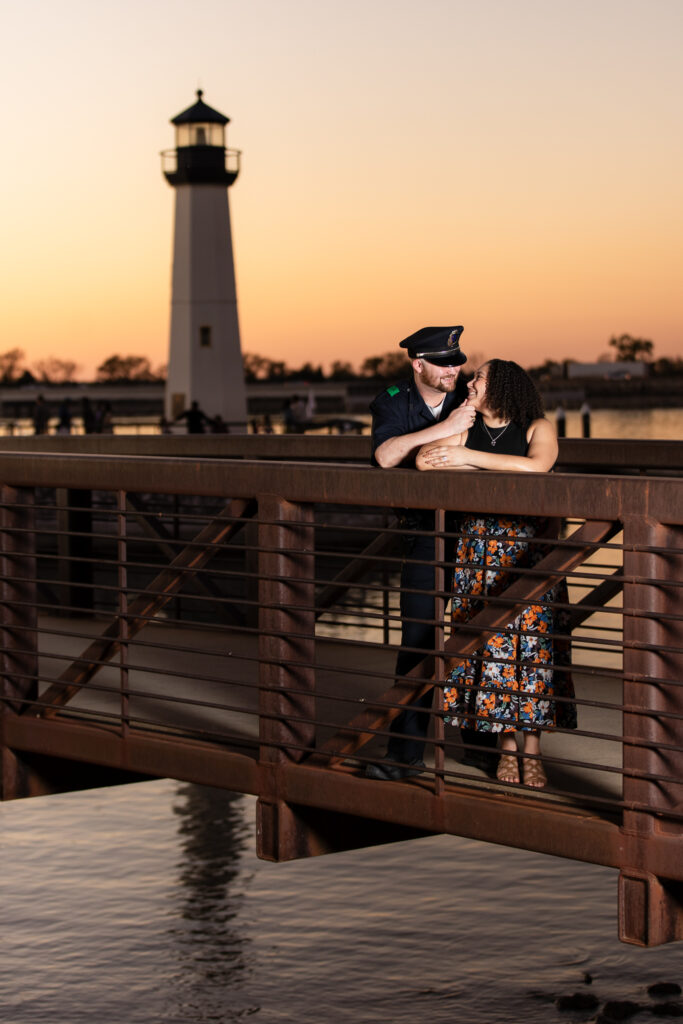 engaged couple standing on bridge cuddling with Rockwall lighthouse behind them at sunset