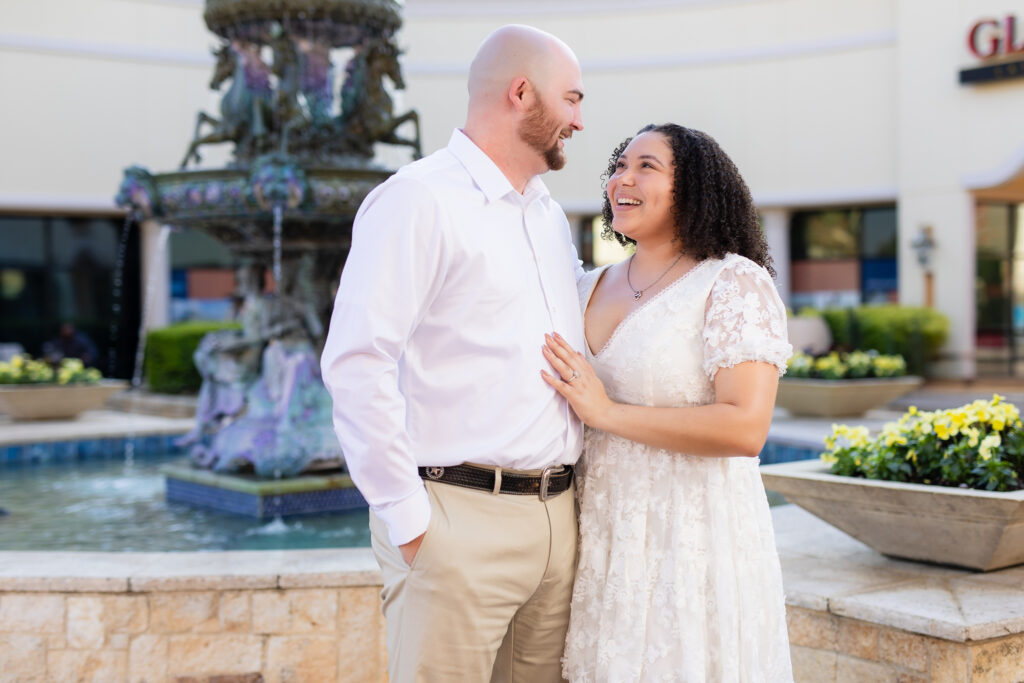 engaged couple wearing all white smiling at each other by Rockwall fountain