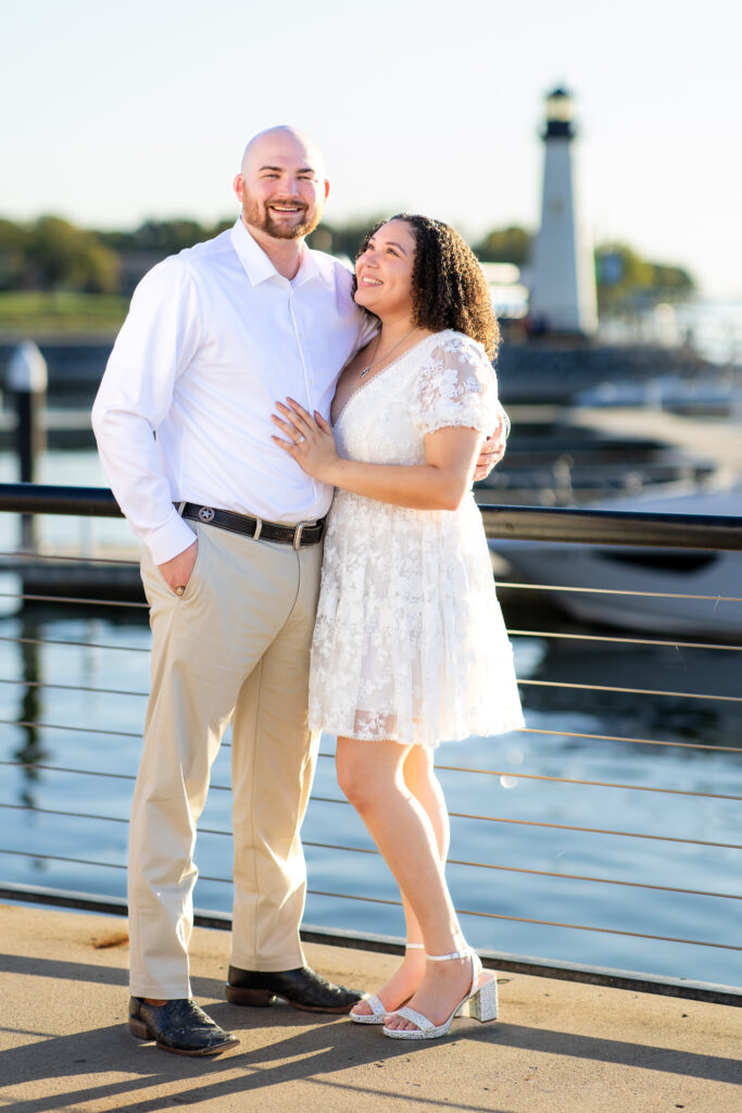 engaged woman looking lovingly up at fiancé by lake with lighthouse in the background