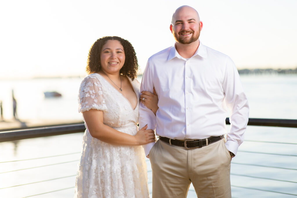 engaged couple in all white linking arms and smiling at camera with the lake behind them