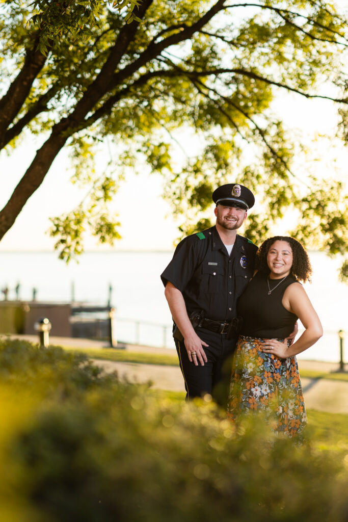 engaged couple smiling at camera under big tree in Rockwall at golden hour during engagement session